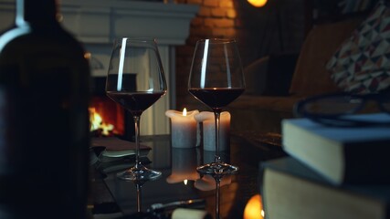 Glass of red wine, book and candle on table in dark room with fireplace. Cozy warm home in winter.