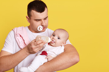 Good looking young man feeding her baby girl isolated over yellow background, handsome young guy with kid's nipple in mouth, Caucasian dad with bottle in hands.