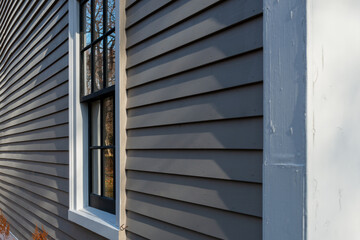 Corner view of a vintage tan coloured house with white trim and black wooden window spacers. The glass in the window is wavy and reflecting the sun. There are orange and red leaves on the ground.