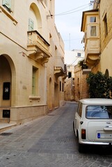 Gozo, Malta: A narrow alley with just enough room for a vintage, cream Innocenti Mini 1000 with Maltese European narrow license plates. Homes with typical Maltese balconies.