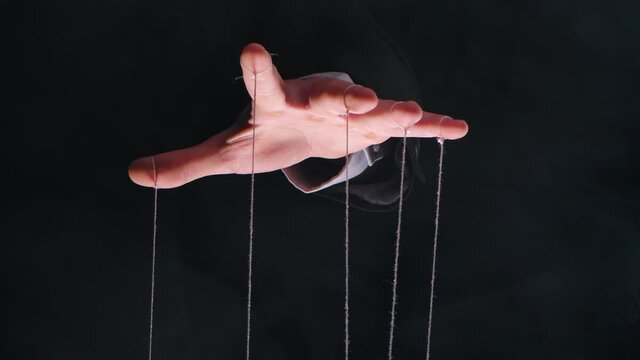 Hand of dictator in a business suit with strings on the fingers to control the puppet. Close up male hand with strings of marionettes on smoke black isolated background. Slow motion.