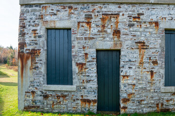 Light grey colored rock wall with brown rust running down in spots. There are two tall blue closed wooden windows and a blue wood narrow door. The building has green grass as a ground covering.