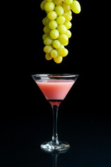 cocktail glass with pink liquid and fresh green grapes with mirrored black background