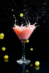 cocktail glass with pink liquid and fresh green grapes, falling and causing a splash with a mirrored black background