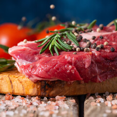 Raw beef meat with spices, tomatoes and rosemary on wooden cutting board o blue background. Image for butcher shop. Macro shot.