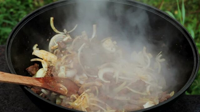 meat with onions is stewed in a large cast-iron caldron over an open fire. Beef is mixed with a wooden spoon and carrots are added to the pan. All ingredients are mixed in the pot.