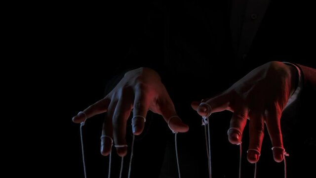 Puppeteer manipulating and pulling strings of marionettes. Male hands close up painted with colored lights on black background. People are toys in hands of dominant businessman dictator. Slow motion.