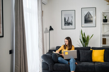 Focused young woman works with a laptop sitting on the couch at home, female student watching a webinar, video courses and taking a notes in a notepad. A female entrepreneur writing plans with a pen