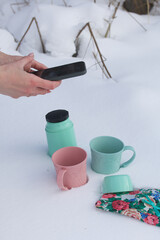Mugs and a thermos are in the snow. Nearby is a colorful napkin. The girl takes pictures of them on a smartphone.