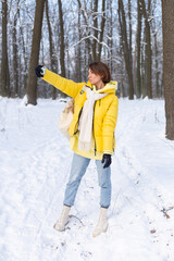 Young beautiful happy cheerful woman in the winter forest video blog, makes a selfie photo against the background of beautiful snow, smiles, dressed in warm clothes, yellow bright jacket