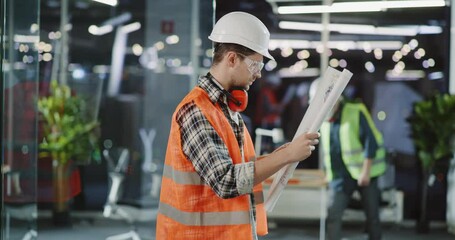 Portrait of middle-aged caucasian hardware worker in protective helmet and vest suit standing at construction site using plan map paper.