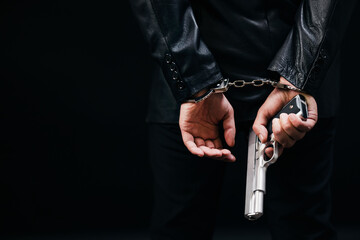 Back view of gangster holding real gun in hands and arrested in handcuffs. Isolated over black studio background. Concept of weapon and criminal.