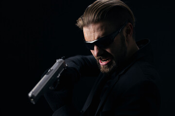 Fototapeta na wymiar Aggressive bearded man in glasses, suit and gloves threatening someone with real gun. Isolated over black background. Concept of danger.