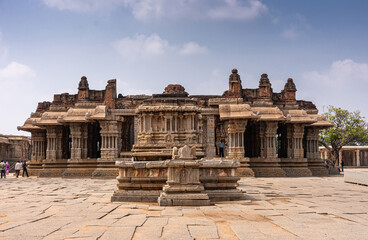 Obraz premium Hampi, Karnataka, India - November 5, 2013: Vijaya Vitthala Temple. Brownstone main sanctuary with mandapam hall seen from central courtyard under blue cloudscape. Clothing adds color.