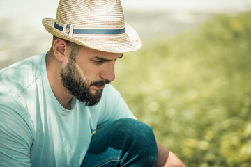 Caucasian serious man with a black beard, a mustache and a hat sits in the nature and looks intently at something down. Dressed in blue jeans and a t-shirt. Summer. Business concept.