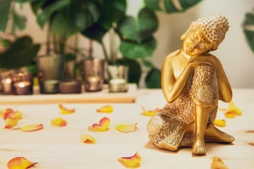 Fotobehang Zen Golden bronze Buddha decorative statuette with flower petals on wooden table with burnig candles, monstera plant background. Meditation ritual, Spirituality, buddhism and religion. Selective focus  © okrasiuk