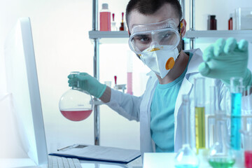 A chemistry laboratory assistant in protective goggles and a mask examines test tubes.