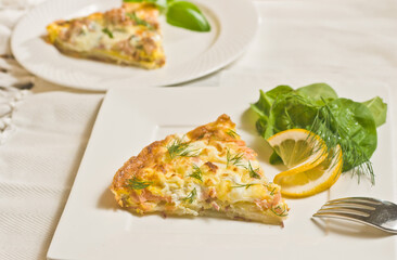 top, front view, medium distance of a slice of warm, baked, homemade, smoked salmon frittata with dill, slice of lemon, on a square, white plate