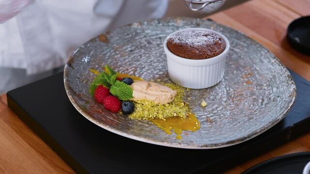 Chef hands sprinkle with powdered sugar through strainer decorating chocolate kulan cake served with ice cream, berries, sauce and pistachio nuts, close up view. Restaurant dessert preparation concept