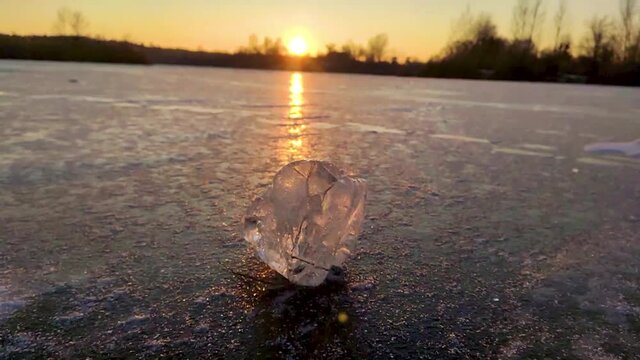 Transparent shining clear icicle slow-motion rotating sparkling on frozen wild lake ice surface in sunset light. Cold winter nature background