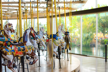 Carousel horses sitting empty at an indoor merry go round carousel in Spokane, Washington with the park and Spokane River blurred in the distance - obrazy, fototapety, plakaty