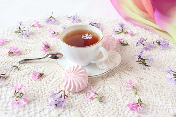 Fototapeta na wymiar White and pink marshmallows on a plate with a cup of tea on a white openwork napkin, delicate flowers nearby, close-up