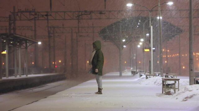Young caucasian woman on empty railway platform in blizzard waiting for a train. Girl missed a train. Female is waiting for the train in bad weather in the evening, strong wind. Bad weather concept