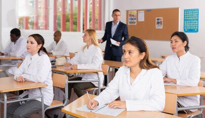 Young woman in white medical coat sitting at desk in classroom attending seminar