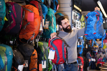 Fototapeta premium Smiling guy customer examining backpacks in sports equipment store