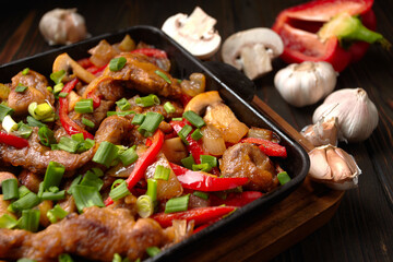 Fried meat with vegetables, green onions, bell peppers and mushrooms, in a frying pan, on a wooden background