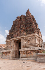 Fototapeta premium Hampi, Karnataka, India - November 5, 2013: Vijaya Vitthala Temple. Red stone gopuram on top of beige stone east entrance gate under blue cloudscape.