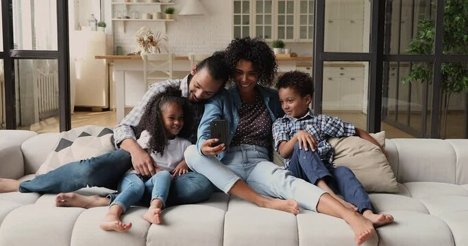 Young African couple their little kids relaxing on comfy cozy couch having fun using mobile app on smartphone, laughing enjoy new cool photo or video editing application. Weekend, modern tech concept