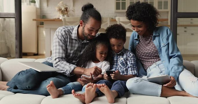 Young African family with siblings kids sit on couch with diverse gadgets, hold tablet laptop and cellphone. Usage of mobile application, bad habits, spend free time use modern tech, overuse concept