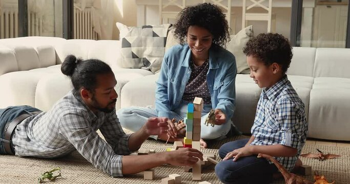 African parents play with 5s cute son dinosaurs and wooden bricks toys resting together on warm floor in cozy living room. Weekend leisure with child, educational games, kid development, fun concept