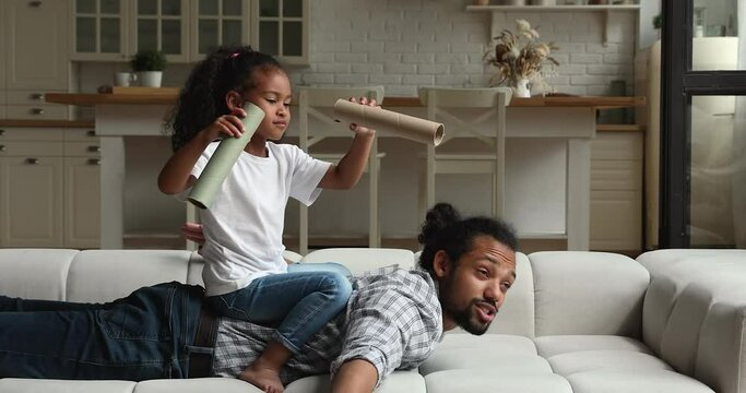 Mixed race family play at home, dad lying on sofa spread his arms like wings of airplane his little daughter sit on daddy back observing into paper tubes imagining it binoculars, funny games concept
