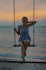 Young happy smiling caucasian woman in dress swinging on a swing standing by the sea on the  beach at sunset