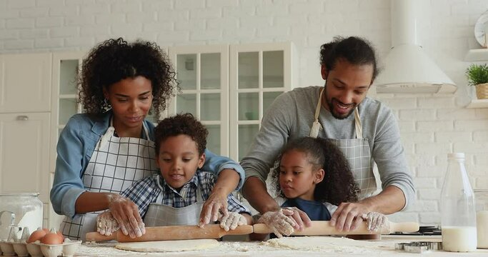 Couple with preschool cute son and daughter preparing dough for cookies, happy African family roll out flattening dough with rolling-pin smiling enjoy process. Homemade healthy food, cookery concept
