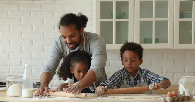 African daddy with preschool 5s son and daughter wearing aprons preparing dough for cookies using rolling-pin flattening dough, making homemade healthy food. Cookery, hobby, happy fatherhood concept