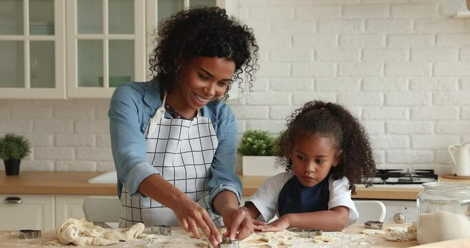 Attractive young African mother and little cute daughter wear aprons cooking together in kitchen, using cookies cutters preparing hand-made biscuits. Hobby, cookery, happy mom teach kid, fun concept