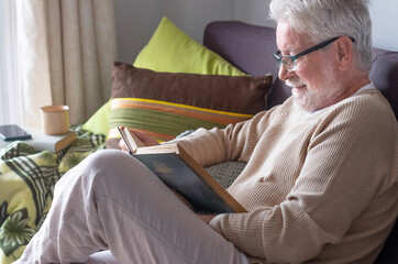 A white-haired elderly man relax at home reading a book on sofa. Bright light from the window. Retired seniors in lockdown due to coronavirus