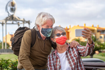 White-haired traveler couple takes a selfie with their mobile phone during the city tour, wearing a surgical mask due to the coronavirus Active retired people enjoying travel and freedom