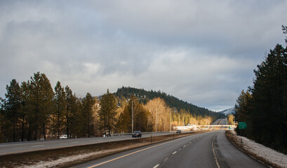Winter landscape -a highway along which cars drive, surrounded by tall trees, on which the rays of the setting sun fall, blue mountains covered with snow are visible in front.Kingston,ID,USA,2-8-2020