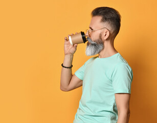 Fototapeta na wymiar Stylish handsome brutal gray-haired bearded man in casual t-shirt and trendy sunglasses drinking morning takeaway cup of coffee. Isolated headshot portrait on orange background. Side view
