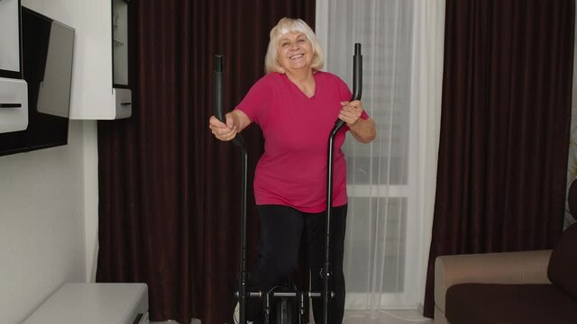 Old grey-haired senior grandmother model exercising workout on orbitrek in room at home. Retro style Caucasian mature elderly woman warming up while sitting on orbitrek indoor. Healthy sport lifestyle