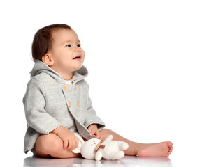 Baby and toy bunny isolated. Barefoot infant child in knitted wear sitting on white floor studio...