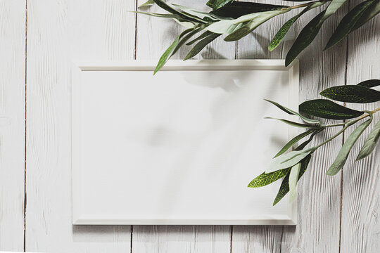 White photo frame with a sprig of eucalyptus on a white wooden table. Minimalistic, gentle mock up