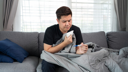 Sick Asian man with flu virus lying in sofa with high fever and measuring body temperature by thermometer and touching his forehead at home