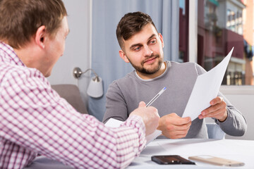 Happy man discussing papers with male colleague while sitting at desk in home interior