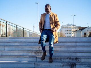 African male wearing jeans with a white turtleneck and beige coat and walking down the stairs