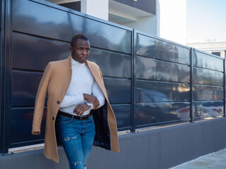 Stylish African male wearing jeans with a white turtleneck and beige coat while leaning on a fence